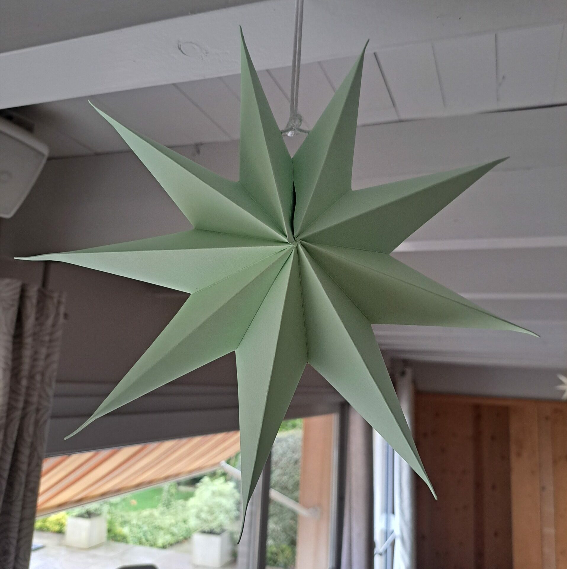Étoile décorative tridimensionnelle en papier de couleur vert sauge, suspendue par un fil à un plafond blanc en bois. Cette décoration de fête en papier plié est utilisée comme suspension intérieure devant une fenêtre.