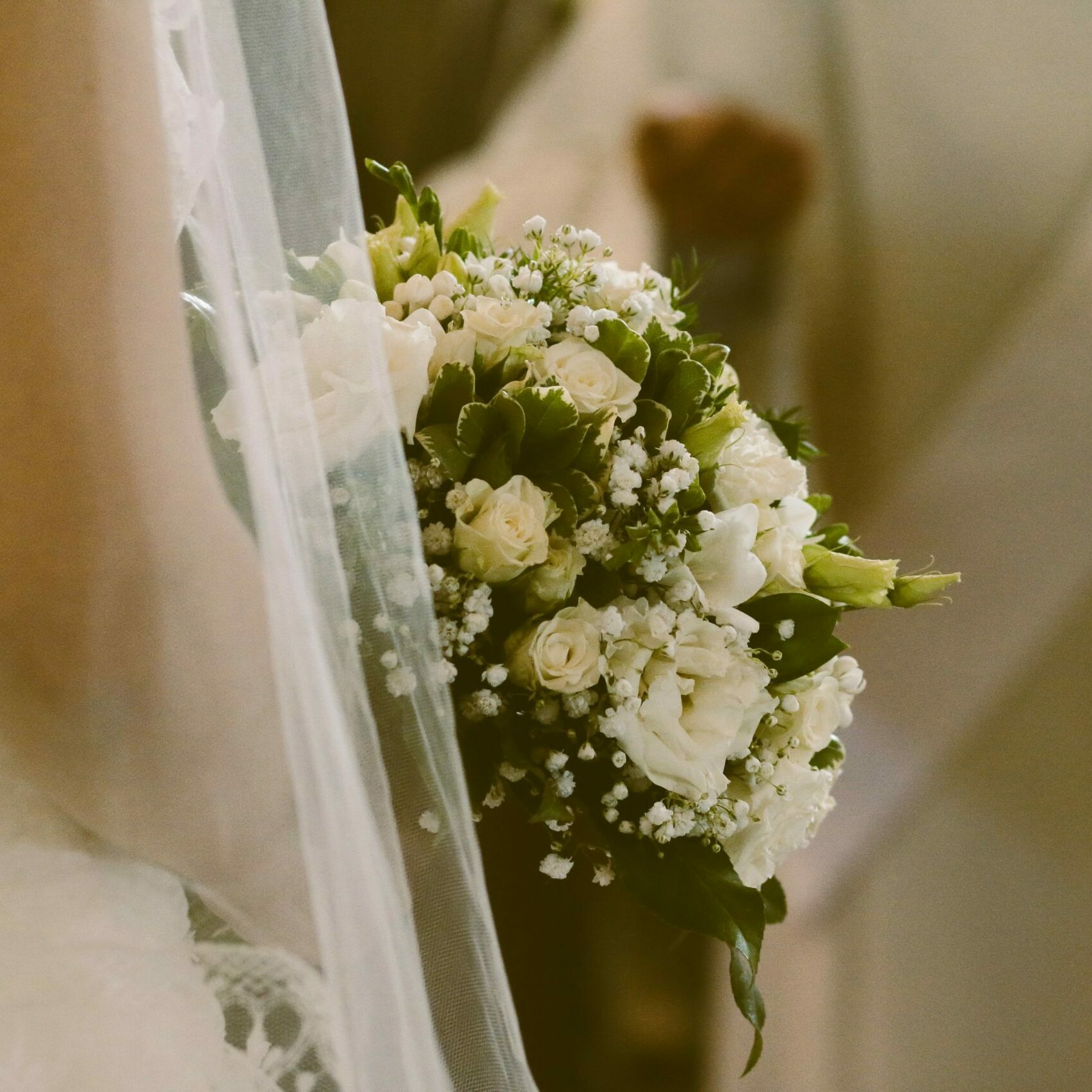 Gros plan sur une mariée tenant son bouquet de mariage. La composition florale inclut des roses de couleur crème, du gypsophile et du feuillage vert, le tout vu à travers le voile de la mariée pour une esthétique douce et vintage.