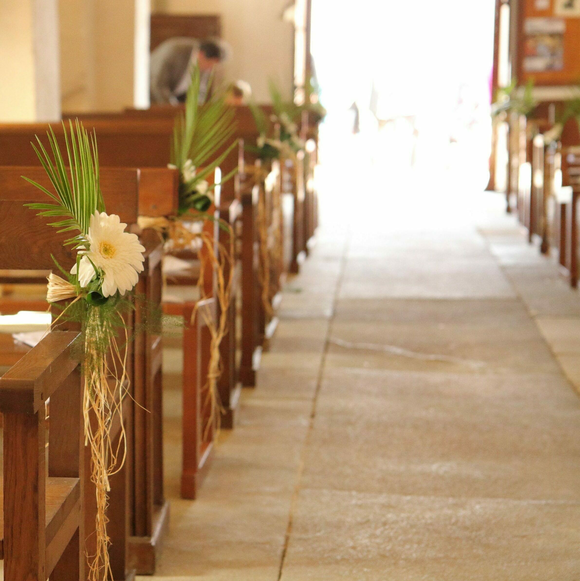 Allée centrale d'une église préparée pour une cérémonie de mariage. Les bancs en bois sont ornés de décorations florales de style tropical, composées d'un gerbera blanc, de feuilles de palmier et de raphia naturel.