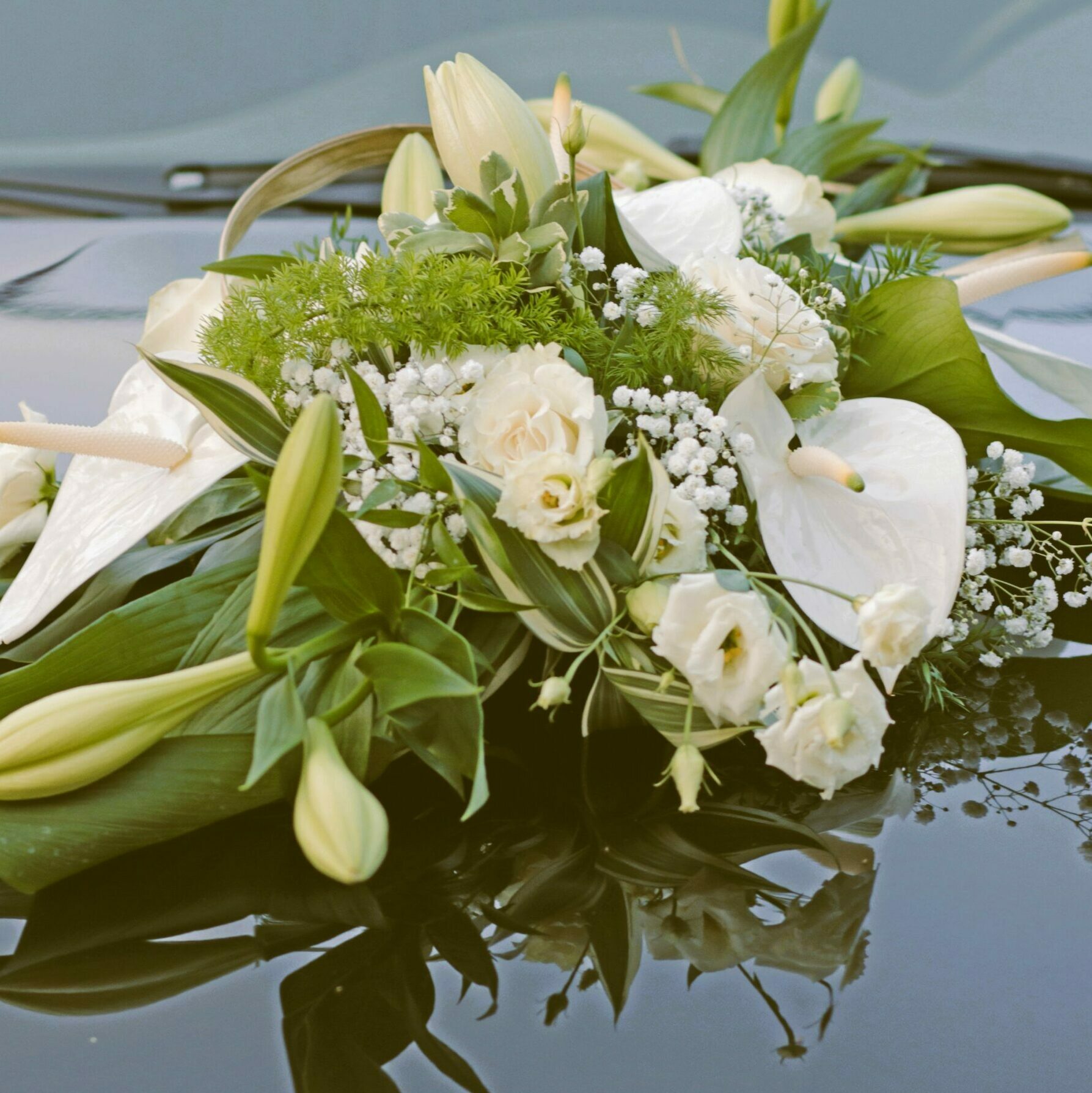 Arrangement floral pour voiture de mariage, posé sur un capot noir réfléchissant. La composition élégante est constituée de fleurs blanches, notamment des lys, des anthuriums, des roses et du gypsophile, ainsi que de feuillage vert.