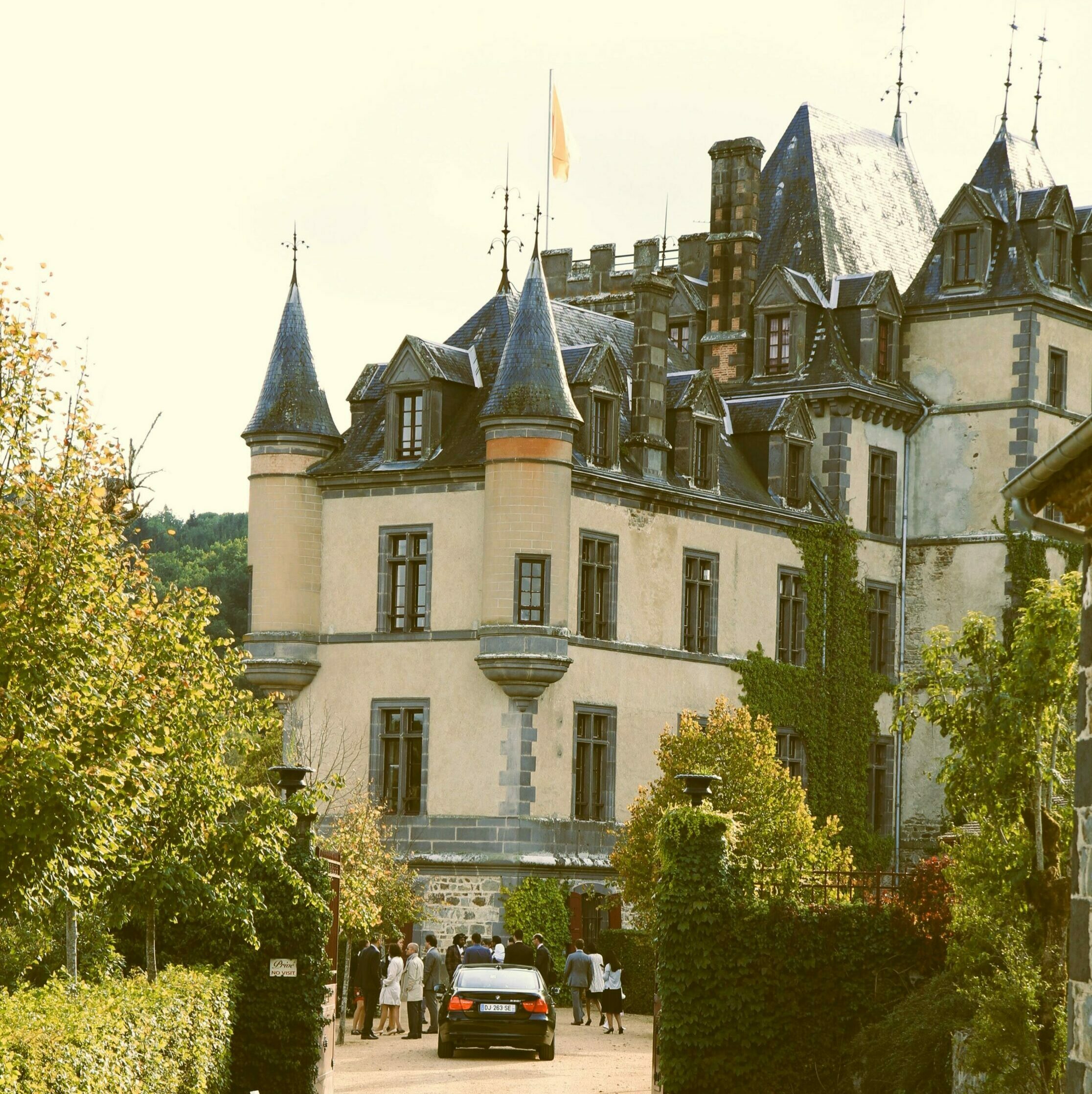 Grand château français utilisé comme lieu de réception de mariage, avec des invités arrivant dans la cour. Le bâtiment possède une architecture classique avec des tourelles, des toits en ardoise et du lierre grimpant sur les murs, le tout dans un cadre verdoyant.
