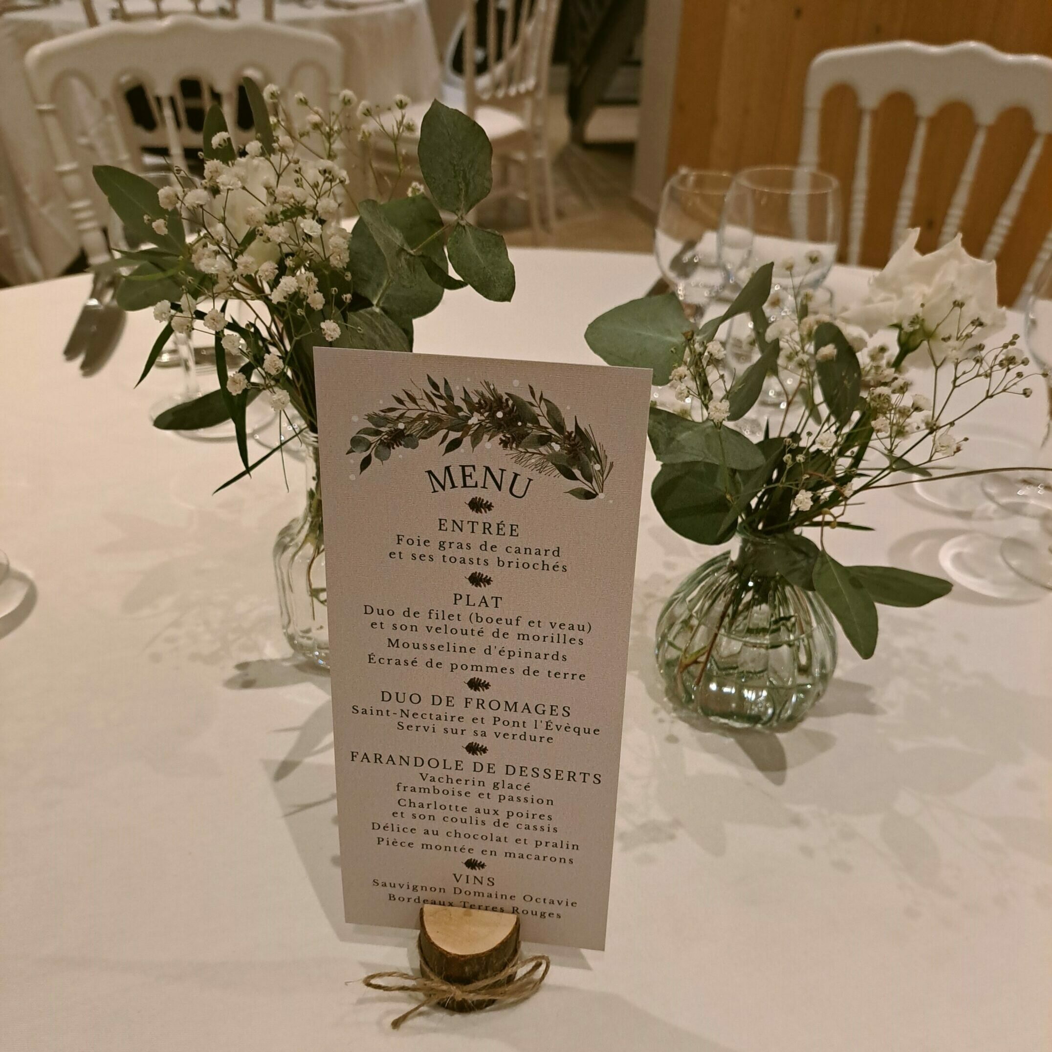Menu de mariage de style rustique, orné de motifs de feuillage, posé sur un support en bois sur une table de réception. La décoration de table comprend des vases en verre avec des compositions florales de gypsophile et d'eucalyptus.