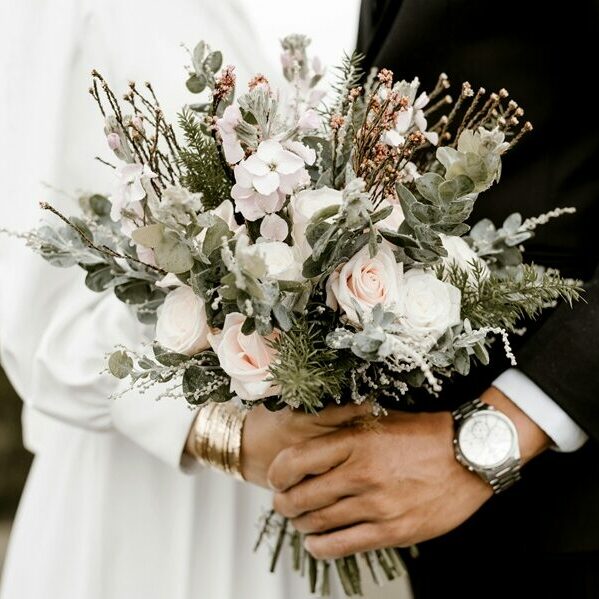 Couple de mariés tenant leur bouquet de mariage, symbole d'une organisation de mariage réussie. Détail sur les mains unies des mariés, la composition florale et les accessoires, illustrant un événement authentique et soigné.