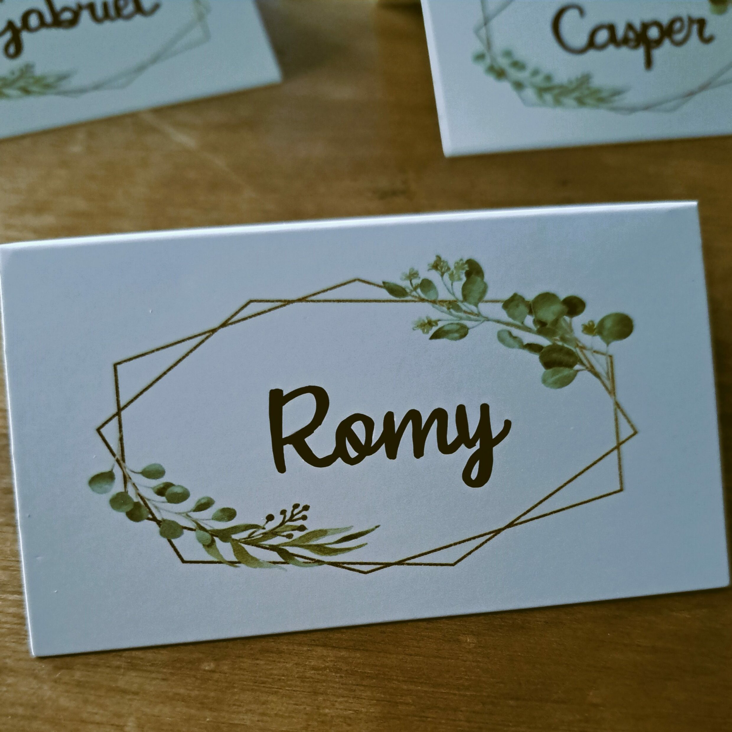 Gros plan sur un marque-place de mariage personnalisé avec le prénom "Romy". Le carton de placement présente un design élégant avec un cadre géométrique doré et des motifs de feuilles d'eucalyptus, posé sur une table en bois.